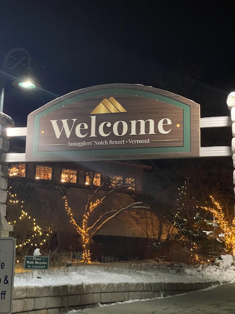 Welcome sign for Smugglers' Notch Resort in Vermont, illuminated at night with surrounding holiday lights.